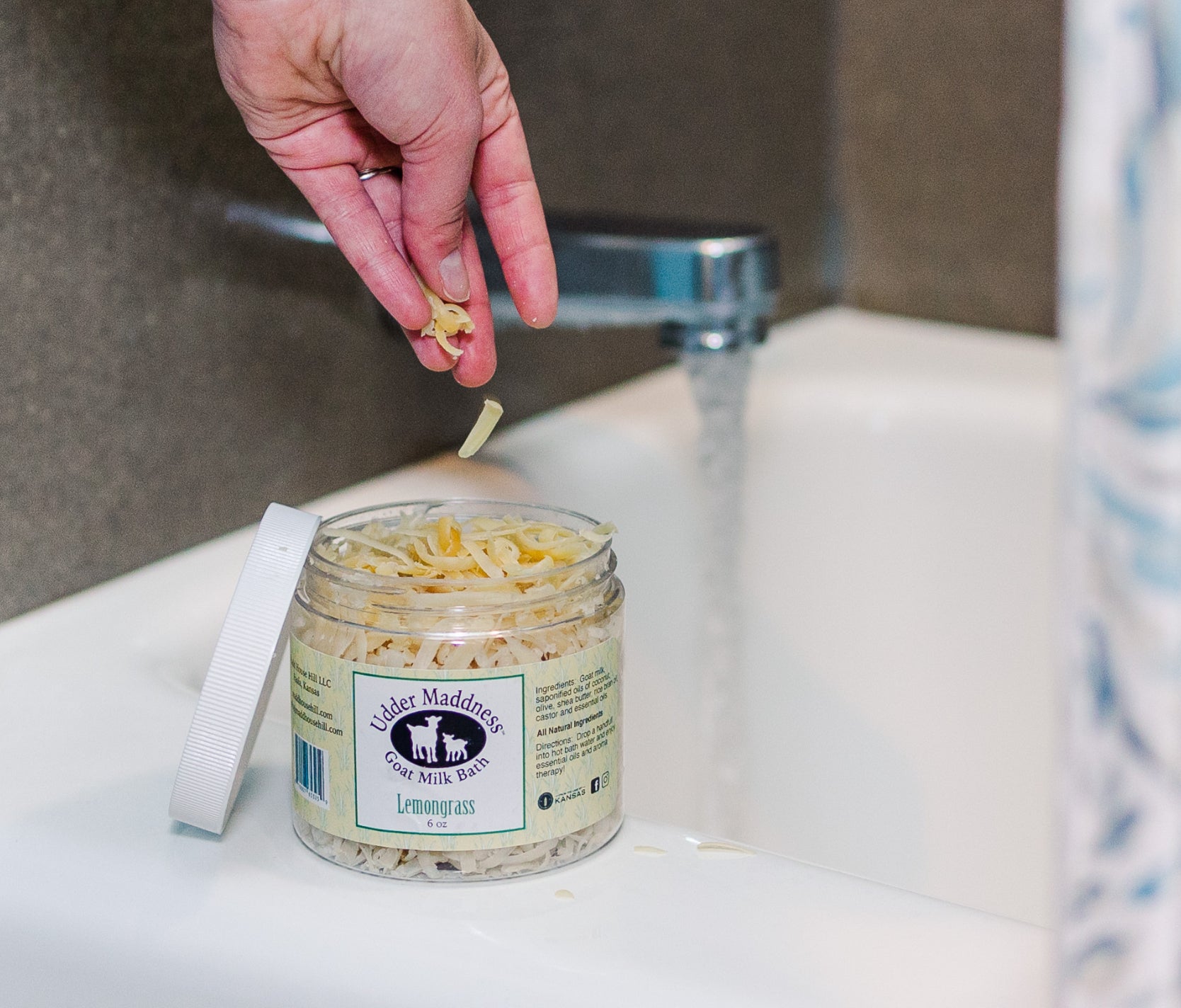 Madd House Hill Udder Maddness Goat Milk Bath Soak — hand dropping milk flakes into jar beside running bath, all-natural skin-softening bath soak