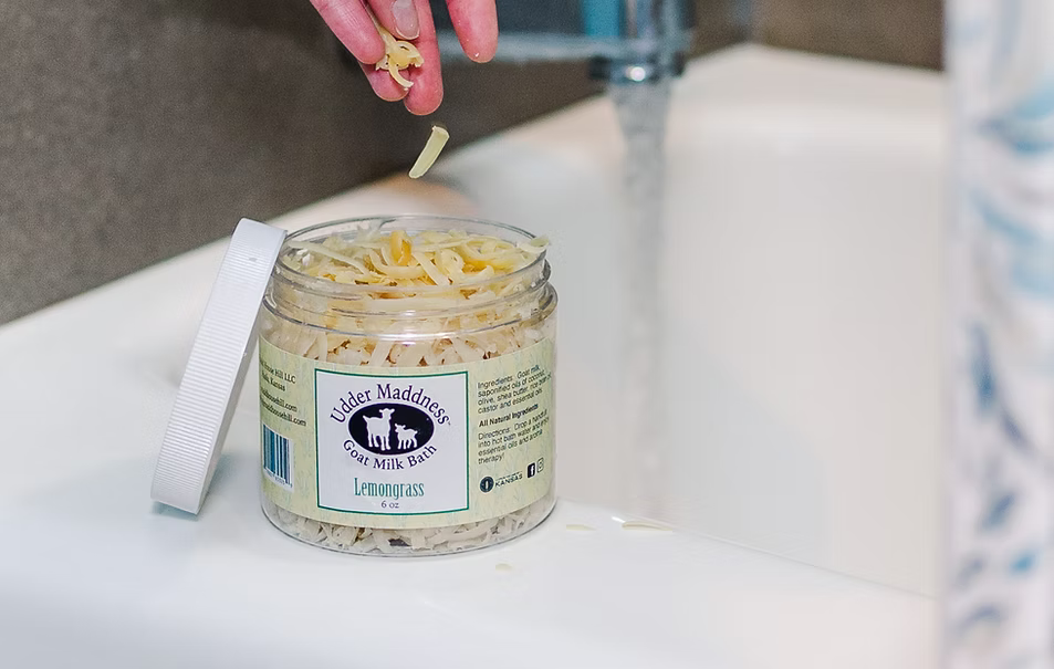 Madd House Hill Udder Maddness Goat Milk Bath Soak — hand dropping milk flakes into jar beside running bath, all-natural skin-softening bath soak